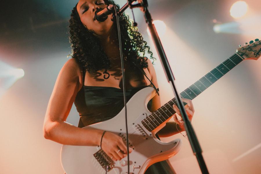 Uwase is playing guitar and singing into a microphone. She is wearing a black dress and has long hair.