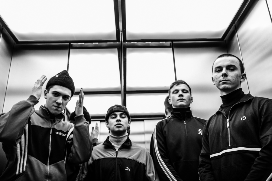 The four bandmembers are standing in an elevator. They are wearing tracksuits.